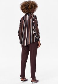 Chemise rayée surdimensionnée en marron, crème et noir ; tissu doux ; manches longues ; portée avec un pantalon ample marron foncé et des chaussures à talons.