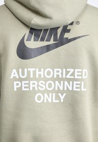 Kapuca olivno zelena s velika črnim logotipom Nike in belim besedilom "LE ZA AVTORIZIRANE OSEBE" na hrbtni strani.