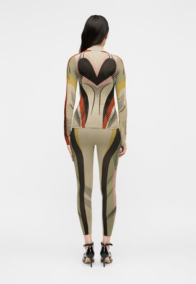 Γυναίκα με μακριά σκούρα μαλλιά που φοράει ένα σετ με μακρυμάνικο τοπ και παντελόνι με μοτίβο σε μπεζ, μαύρους, πορτοκαλί, κίτρινους και ροζ τόνους, με μαύρα τακούνια.