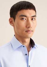 Camicia azzurro chiaro con colletto nero a fantasia, chiusura con un solo bottone e bottoni neri a contrasto. Tessuto dalla texture liscia.