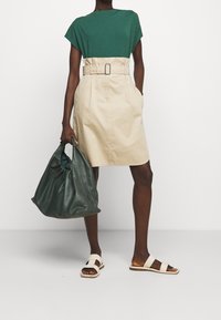 Groene shirt met korte mouwen gecombineerd met een beige rok met riem, houdt een donkergroene leren tas vast en draagt crèmekleurige slide-sandalen.