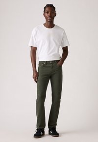 Jeans in cotone verde e una t-shirt bianca a collo rotondo, abbinati a sneaker nere. L'outfit presenta un taglio rilassato e dettagli minimal.