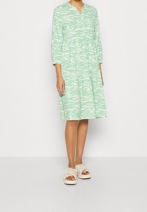 Mujer que lleva un vestido verde menta hasta la rodilla con patrones abstractos blancos y mangas tres cuartos, combinado con sandalias beige con fruncidos.
