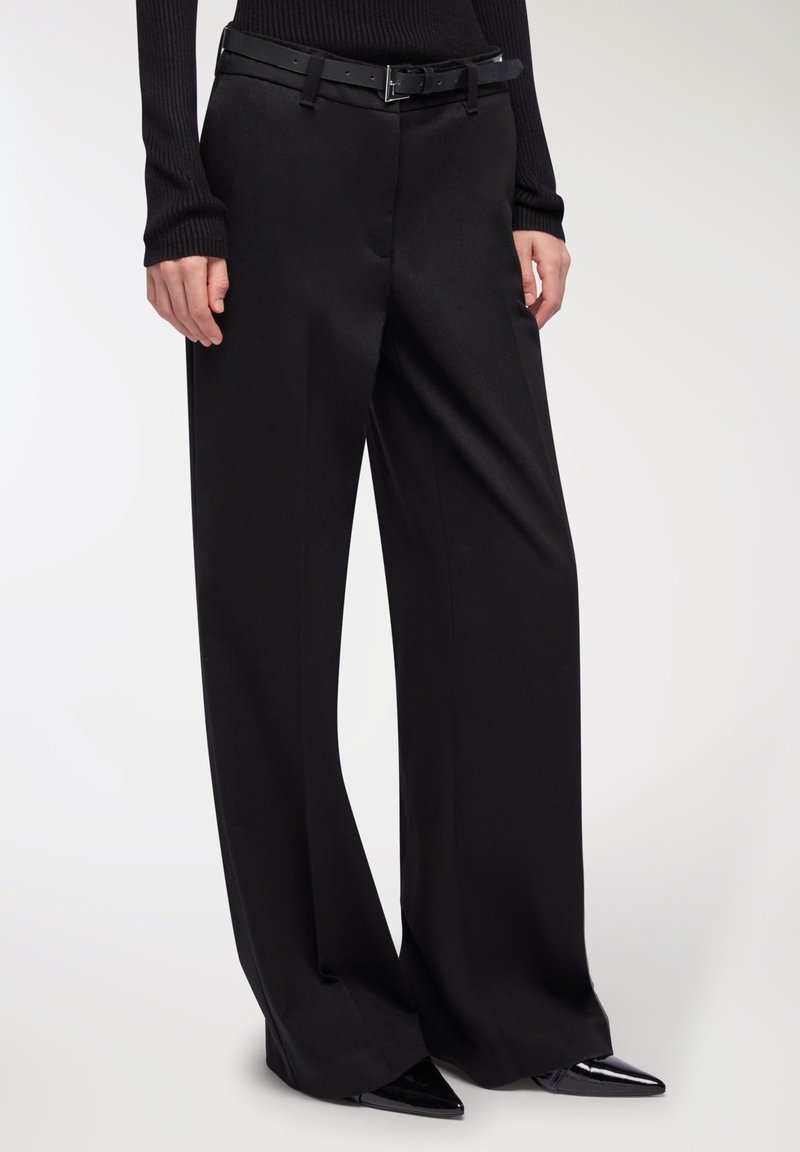 Pantaloni larghi neri realizzati in un tessuto liscio, con una vita aderente e una cintura nera, accompagnati da scarpe nere appuntite e affilate.