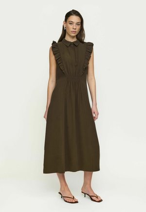 Kvinde stående iført en ærmeløs mørkebrun midi-kjole med flæsedetaljer på skuldrene og sorte hælede sandaler foran en ensfarvet baggrund.