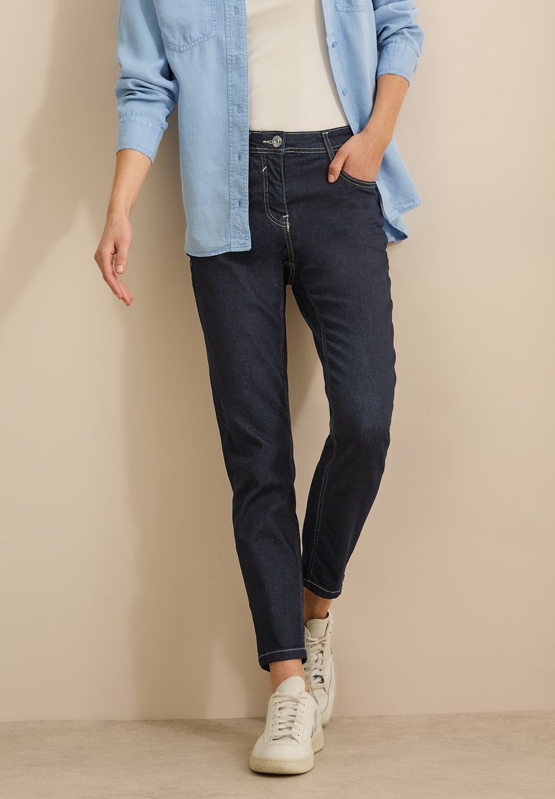Jean bleu foncé avec une coupe slim, featuring une taille mi-haute, cinq poches et des coutures jaunes contrastées, associé à des baskets blanches.