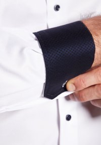 Camicia bianca con un polsino testurizzato di un blu scuro. Il polsino presenta un piccolo motivo a quadretti e cuciture a contrasto. Bottone nero visibile.
