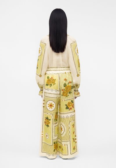 Γυναίκα με μακριά σκοτεινά μαλλιά που φοράει μπεζ παντελόνια με φαρδιά μπατζάκια και ανάλογο μπλουζάκι με μακριά μανίκια με κίτρινα λουλουδάτα και γεωμετρικά σχέδια, από πίσω.