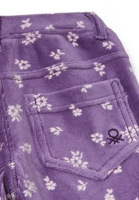 Tissu en velours côtelé violet avec un motif floral subtil. Présente une poche arrière avec un symbole brodé. Texture douce, détails de couture distincts.