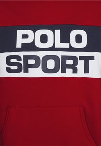 Felpa rossa con strisce orizzontali blu e bianche. Presenta un grande testo "POLO" e "SPORT" in colori a contrasto e in grassetto. Include tasca a marsupio.