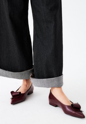 Bailarinas de cuero burdeos con un detalle de lazo en la punta y un tacón cuadrado bajo, combinadas con pantalones de mezclilla oscuros y remangados.