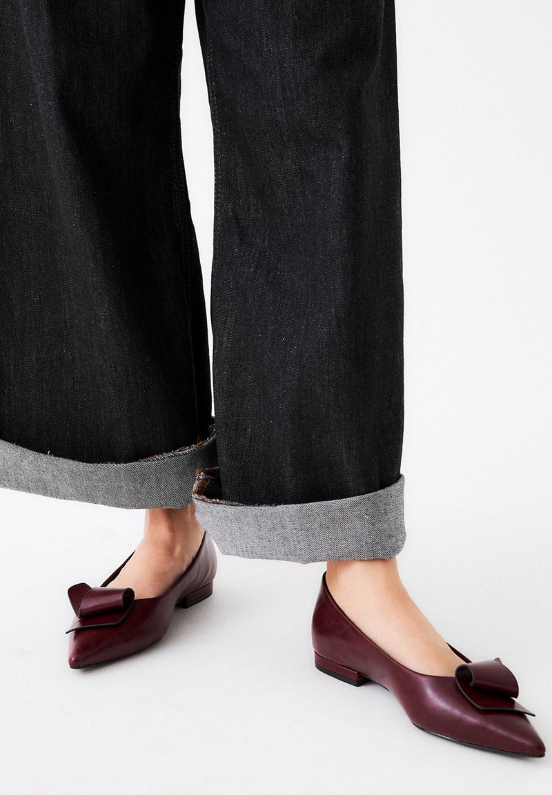 Bailarinas de cuero burdeos con un detalle de lazo en la punta y un tacón cuadrado bajo, combinadas con pantalones de mezclilla oscuros y remangados.