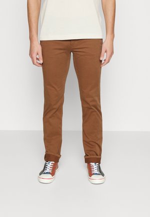 Person iført opsmøgede brune bukser og flerfarvede sneakers, stående mod en ensfarvet hvid baggrund.