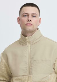 Giacca beige con zip, con un lato sinistro in fleece testurizzato e un lato destro in tessuto liscio. Presenta un colletto alto e una tasca sul petto.