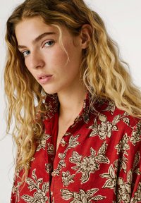 Rode button-up shirt met een bloemenpatroon in beige en donkergroen. De stof lijkt lichtgewicht met een iets gestructureerde afwerking.