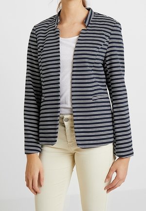 Femme portant un blazer à rayures horizontales bleu marine et blanches, avec un haut blanc et un pantalon beige, debout devant un fond uni.