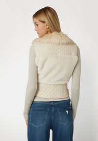 Bežinė siuvinėta liemenė su dirbtinio kailio apykakle, tekstūruotu audiniu ir banguotomis rankovėmis. Iš galo matosi lygus, minkštas audinys.