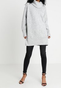 Harmaan ribbed neulemekko, jossa on kauluksellinen kaulus, pitkät hihat ja sivuaukot, yhdistettynä mustiin slim fit -farkkuihin ja mustiin remmikenkiin.