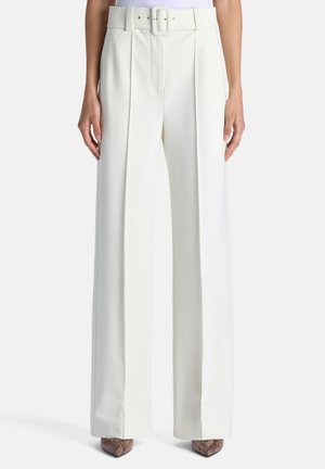 Pantalon blanc taille haute à jambes larges avec plis à l'avant et ceinture assortie, porté avec des chaussures marron pointues.