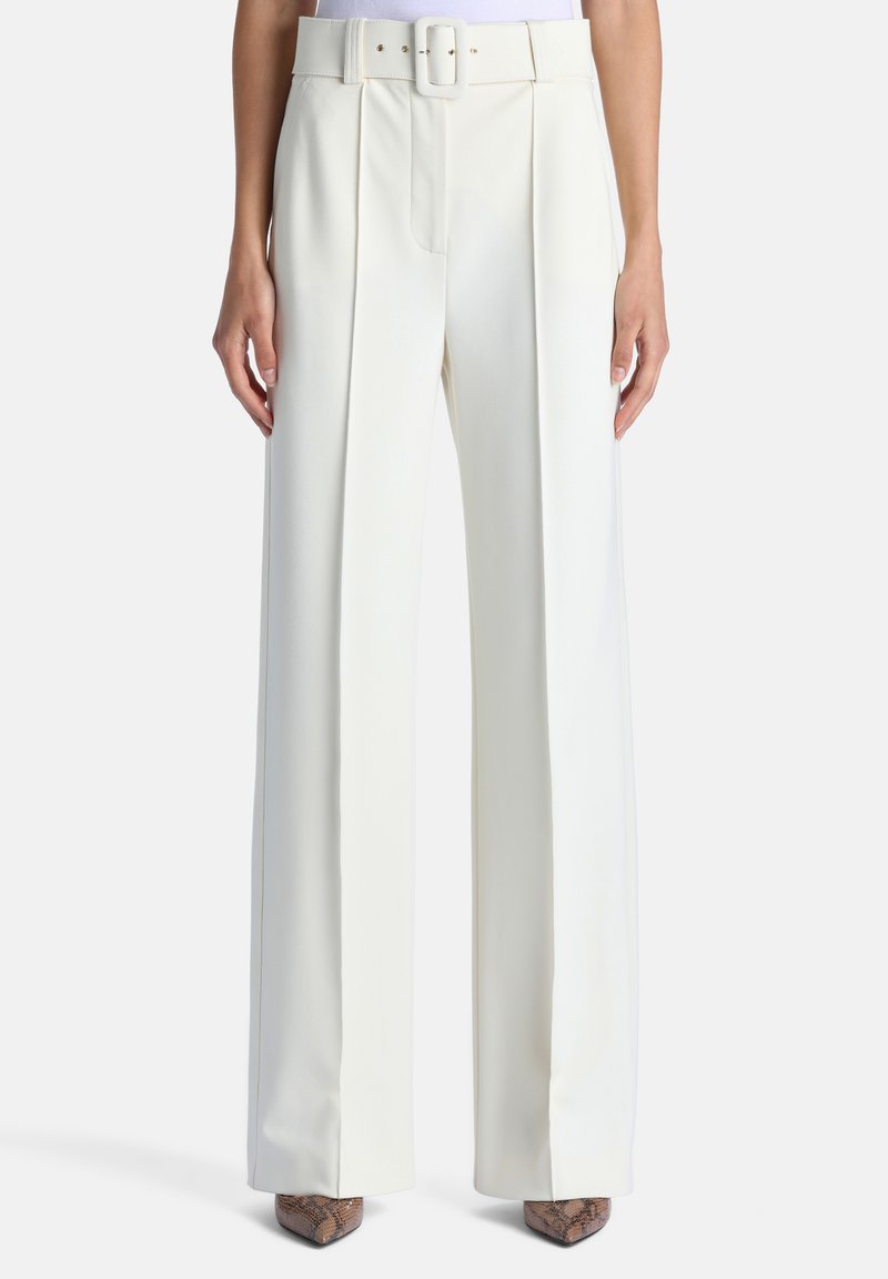 Pantalon blanc taille haute à jambes larges avec plis à l'avant et ceinture assortie, porté avec des chaussures marron pointues.