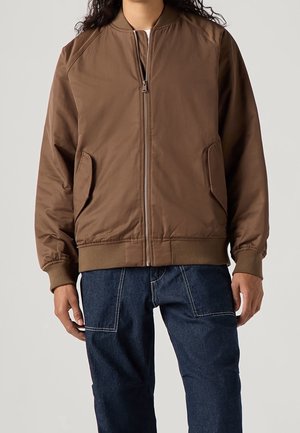 Person trägt eine braune Bomberjacke mit Reißverschluss und Klappentaschen sowie dunkelblaue Jeans mit großen Fronttaschen.
