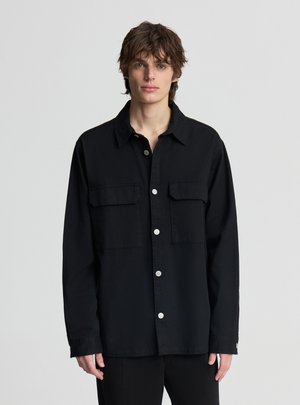 Giovane uomo con capelli castani scompigliati, indossa una camicia nera con bottoni e due tasche sul petto, in piedi davanti a uno sfondo grigio chiaro.