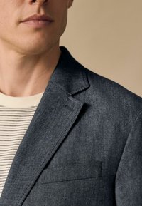 Tmavošedý blazer s jemnou štruktúrou, so zárezmi na klopách a vreckom na ľavej hrudi, nosený cez pruhovanú svetlú košeľu.