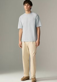 Lyseblå kortærmet t-shirt med brystlomme, båret med beige bukser og brune sko, stående foran en neutral baggrund.