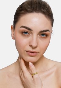 Bague en chaîne dorée sur une main, avec des maillons entrelacés. Peau lisse et maquillage naturel avec des yeux bleus et un arrière-plan neutre.