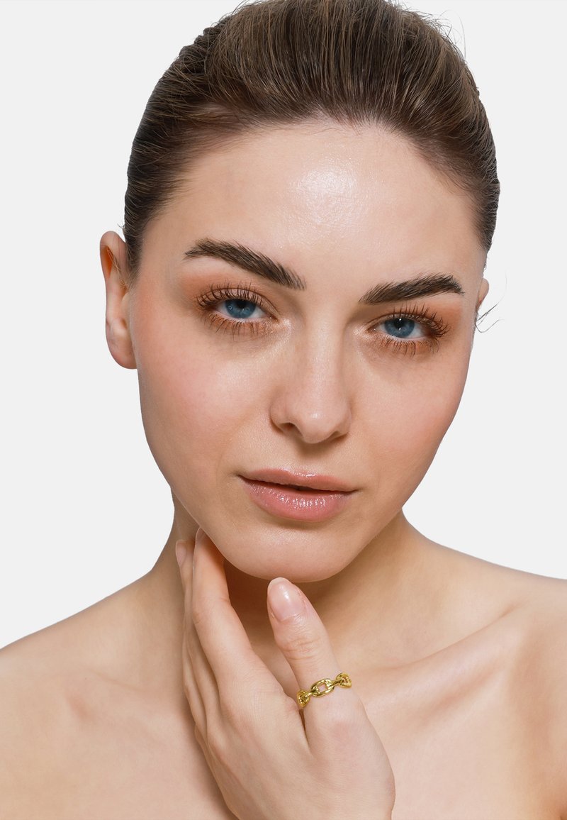 Bague en chaîne dorée sur une main, avec des maillons entrelacés. Peau lisse et maquillage naturel avec des yeux bleus et un arrière-plan neutre.