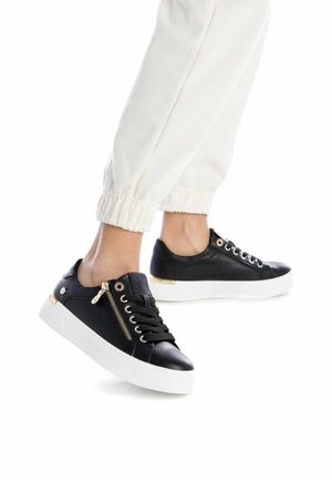 Zapatillas de cuero negro con una suela de goma blanca, con detalles en cremallera dorada, cordones negros y una textura suave. Acentuadas con herrajes dorados.