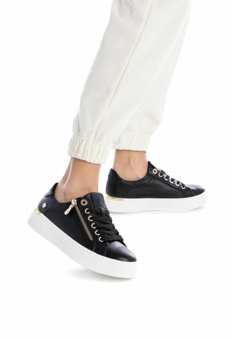 Zapatillas de cuero negro con una suela de goma blanca, con detalles en cremallera dorada, cordones negros y una textura suave. Acentuadas con herrajes dorados.