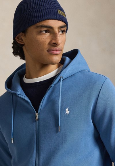 Ανοιχτό μπλε φούτερ με φερμουάρ και λευκό λογότυπο στο στήθος, με κουκούλα με κορδόνια και απαλό ύφασμα. Συνδυασμένο με σκουρό μπλε πλεκτό σκουφάκι.