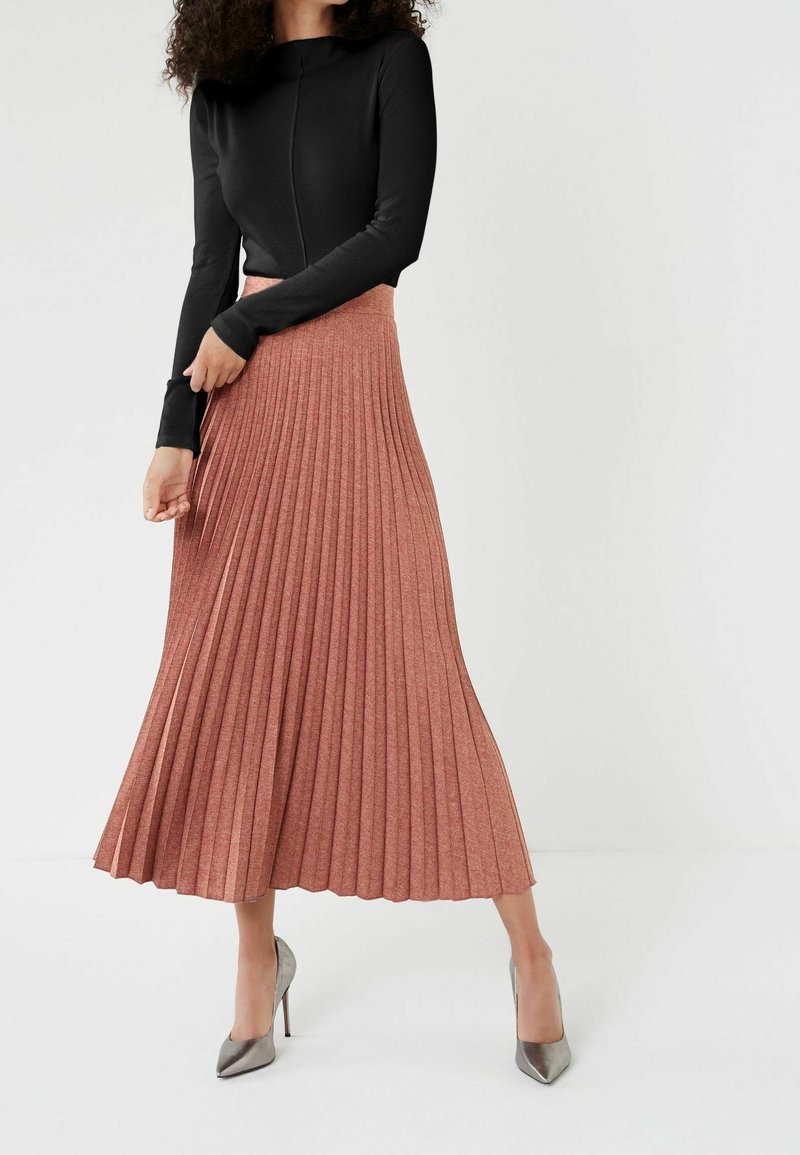 Femme portant un haut noir à manches longues, une jupe plissée taille haute couleur rouille et des chaussures à talons hauts argentées, debout les jambes croisées.