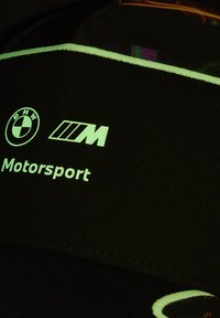 Schwarze Mütze mit BMW-Logo, "M"-Insignie und "Motorsport"-Text. Der Text ist weiß mit einem reflektierenden Finish und liegt auf einem gerippten Stoffhintergrund.
