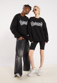 Sorte hettegensere med "Visionary" i sølvfarge, kombinert med slitte sorte jeans og hvite høye sneakers. Hvit bakgrunn.