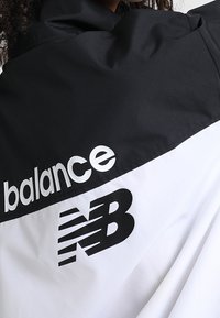 Czarna i biała kurtka przeciwwiatrowa z napisem "balance" oraz logo "NB" w czarnym nadruku. Posiada gładką fakturę i sportowy design.