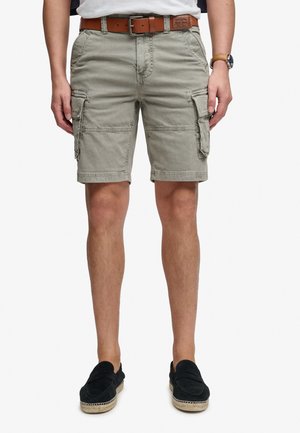 Man draagt beige cargoshort, bruine leren riem, zwarte instapschoenen en een polshorloge, staand tegen een witte achtergrond.