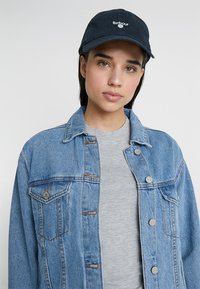 Leren spijkerjas in lichtblauw met zilveren knopen en borstzakken, gelaagd over een grijs shirt, gecombineerd met een marineblauwe honkbalpet met een wit logo.