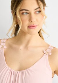 Top senza maniche di colore rosa, caratterizzato da un tessuto texturizzato, scollatura arricciata e applicazioni floreali sulle spalline per un tocco di dettaglio in più.