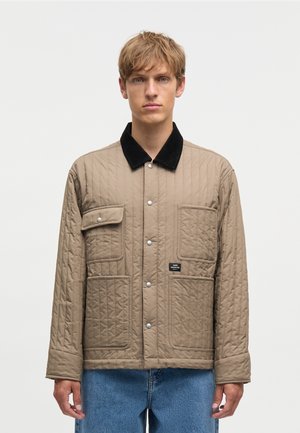 Ung mand iført en beige quiltet jakke med sort krave, trykknapper, flere forlommer og blå jeans mod en ensfarvet baggrund.