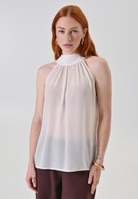 Top trasparente senza maniche di un leggero beige con colletto alto, dettagli arricciati e una silhouette fluida, abbinato a pantaloni marrone scuro.