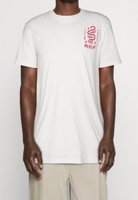 Bílý bavlněný tričko s krátkým rukávem. Na levé straně hrudi je červený grafický design se hadem a textem. Měkká textura.