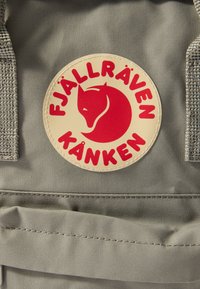 Harmaa reppu, jossa on pyöreä, beige kangasmerkki, jossa on punainen ketunlogo ja teksti "FJÄLLRÄVEN KÅNKEN" paksuilla kirjaimilla. Kankaan pintakuviointi.