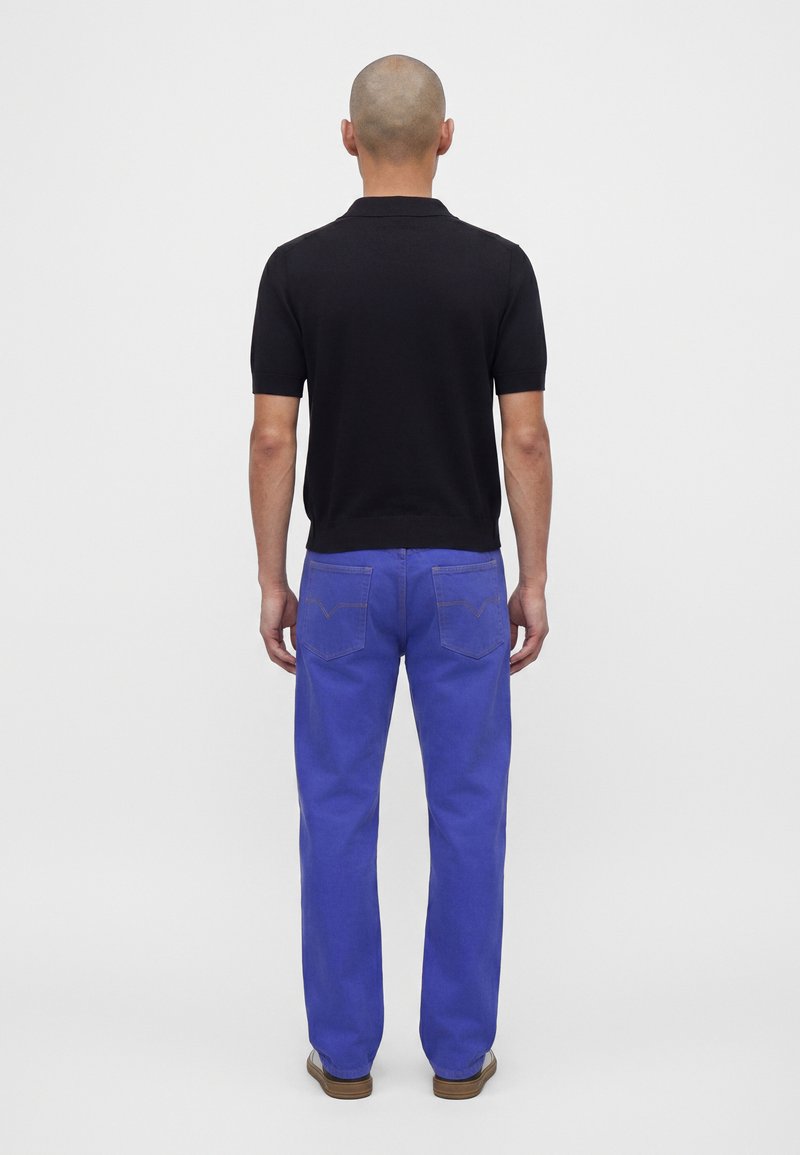 Homme à la tête rasée portant une chemise noire à manches courtes, un pantalon bleu vif et des chaussures marron, debout de dos sur un fond uni.
