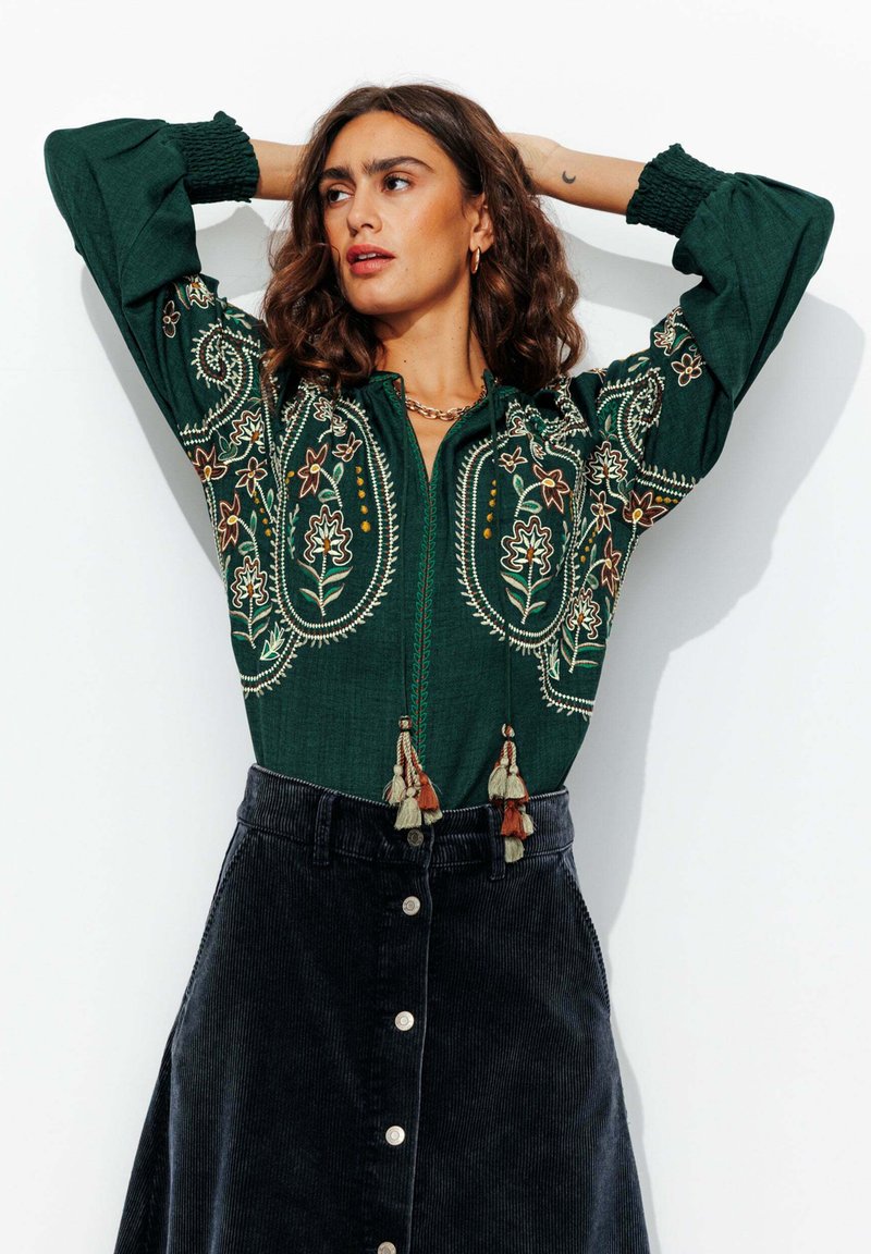 Femme aux cheveux bouclés portant un chemisier vert brodé avec des glands et une jupe en velours côtelé noire à boutons, posant les mains derrière la tête.