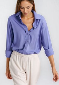 Koszula z długim rękawem na guziki w jasnofioletowym materiale o luźnym kroju, z rozłożystym kołnierzem i podwiniętymi mankietami. W zestawie z beżowymi spodniami.