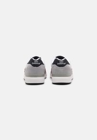 Pár šedobílých tenisek New Balance zobrazených zezadu, s černými patními poutky s logem značky na jednolitém pozadí.