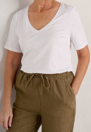 Bejaarde persoon draagt een wit T-shirt met V-hals en korte mouwen, ingestopt in olijfgroene linnen broek met elastische taille en trekkoord.