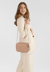 Giovane donna in completo beige che tiene una borsa a tracolla trapuntata beige con tracolla a catena, sorridente e con lo sguardo rivolto di lato su uno sfondo semplice.